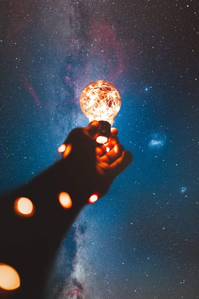 A hand holds a glowing bulb into the Milky Way night sky, creating a magical cosmic connection.